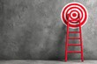 © Adikx - A red ladder leans against a large red and white target painted on a textured grey wall.  The scene evokes ambition and achieving goals