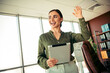 © deagreez - Cheerful businesswoman waving hello while holding a tablet in a modern office workspace