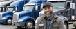 © lililia - Happy African American truck driver stands with arms crossed in front of blue trucks, smiling confidently in a parking lot