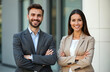 © miss irine - Young business partners, man and woman, pose outside office. Smiling confident business people stand with crossed arms. Successful team ready for partnership and corporate events.