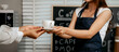 © Thitaporn - A young Asian female barista in a striped apron and orange shirt stands in a cozy modern café, smiling warmly as she hands a freshly made coffee cup to a woman customer.