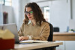 © insta_photos - Smiling female student using computer technology learning online, doing web research. Young happy professional business woman worker employee sitting at desk working on laptop in corporate office.