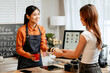 © Thitaporn - A young Asian female barista in modern cafe assists woman customer ordering latte, fresh coffee beans, dark roast, croissant, completing the purchase with mobile payment at the POS using QR code.