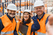 © Koldo_Studio - Smiling warehouse workers team taking a selfie, wearing safety vests and helmets, celebrating successful teamwork in a logistics center
