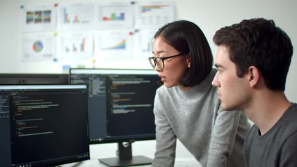 Two Young Professionals Collaborate on Coding at Monitors with Charts in White Background and Gray Outfits Bright Lighting