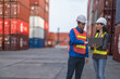 © ultramansk - Two logistics workers man and woman discuss operations at a busy shipping container yard.