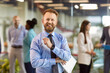 © Studio Romantic - Portrait of a young happy smiling confident bearded business man in formal clothes looking cheerful at camera with team of company employees working on background in office. Business portrait concept