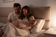 © ilyaska - Expecting parents tenderly touching pregnant belly while sitting together in bed, enjoying intimate moment in soft evening light