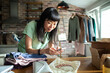 © Marko Geber - Woman photographing handmade jewelry at home for online store surrounded by clothes and packing materials