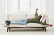 © SHOTPRIME STUDIO - A joyful Black man relaxing on a couch, wearing a green sweater and blue jeans, embodying comfort and happiness in a bright, airy living space
