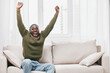 © SHOTPRIME STUDIO - Happy Black man celebrating success in a cozy living room, wearing a green sweater, embodying joy and excitement with a bright atmosphere