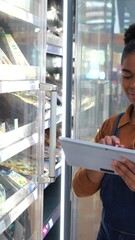 Professional young african american woman using digital tablet for inventory management, checking refrigerated products in modern grocery store with efficiency and precision