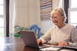 © luciano - Smiling mature woman sitting at table with laptop and books working or studying. Attractive senior woman in eyeglasses enjoying teaching activity and learning using new technology