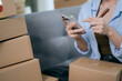 © Dara-Sroi - An Asian female entrepreneur prepares parcels and checks online orders from her home using her laptop and smartphone, reflecting the concept of online shopping and starting a SME business.