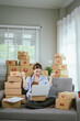 © Dara-Sroi - An Asian female entrepreneur prepares parcels and checks online orders from her home using her laptop and smartphone, reflecting the concept of online shopping and starting a SME business.