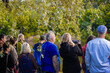 © Valters - A diverse group of people stands near a calm forest lake, watching an event or view, with one person filming the scene using a smartphone under leafy tree branches.