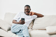 © SHOTPRIME STUDIO - A smiling Black man in casual attire enjoying his smartphone on a comfortable sofa, conveying relaxation and modern lifestyle