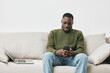 © SHOTPRIME STUDIO - Young Black man sitting on a sofa, smiling while using a smartphone, wearing a green sweater and jeans in a modern, bright living space