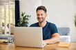 © baranq - Smiling man using laptop at home sitting at desk