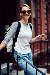 Wall Mural - Half length portrait of cheerful young woman in sunglasses standing on stairs holding smartphone,positive hipster girl in casual wear looking at camera sending message via mobile phone on street