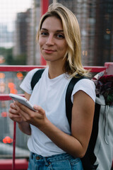 Wall Mural - Half length portrait of young female tourist carrying backpack with flowers sightseeing  in city using mobile phone application for navigate, hipster girl standing on urban bridge with smartphone