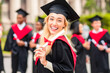 © Prostock-studio - Cute blonde lady student having graduation party, wearing graduation robe and hat, holding diploma and smiling at camera, posing over international group of graduates, panorama