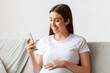 © Prostock-studio - Happy pregnant millennial woman using smartphone at home, browsing pregnancy app while sitting on couch in living room, smiling expectant lady looking at phone screen and touching belly, free space