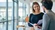 © mangolovemom - Two professionals engage in a friendly conversation in a modern office with large windows and natural light.