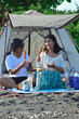 © agratitudesign - Two smiling teenage girls enjoy sausage skewers and conversation on a mat under a beach canopy, in front of a tent surrounded by tropical greenery.