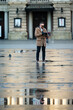 © Niko_Dali - Urban solitude captured in a quiet square where reflections dance on rain-soaked pavement as a man engrosses in his phone amid the chirping of curious pigeons