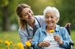 © Vadym - Smiling nurse caregiver in uniform holds flower with senior woman in wheelchair. Elderly lady enjoys sunny day, relaxing in park. Healthcare, support, nursing home concept.