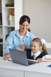 © Evgeniya Grande - Vertical lifestyle portrait young mother sits at the table with daughter and helps little schoolgirl do her online homework on the computer at home. Woman tutor teaches a girl lessons on a laptop