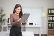 © MINAE - Young Asian businesswoman smiling while using a digital tablet in a modern office, embodying the synergy of technology and business in a bright, organized workspace