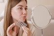 © st.kolesnikov - Teenager with skin blemishes holds acne patches by her face in front of a mirror, showing modern skincare and selfcare treatment.