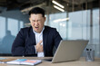 © Tetiana - Asian young male businessman sitting in office at desk and holding his hand on his chest, feeling severe heart pain