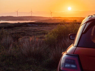 Wall Mural - A red car is parked in a field with wind turbines and a warm sunrise in the background. The car is facing the sun, and the sky is orange and pink. The scene is peaceful and serene. Early bird concept