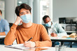 © Prostock-studio - Modern Education Concept. Portrait of pensive asian student sitting at table in classroom at college, wearing surgical medical mask, writing in notebook and thinking, looking aside. Banner, free space