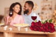 © Prostock-studio - Romance And Bonding. Selective focus on bunch of red roses, bouquet of flowers lying on table. African american couple spending time together, having dinner, drinking wine in blurred background