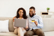 © Prostock-studio - Online Wallet And Banking Concept. Happy young african american family of two people using laptop and making purchases via internet store, paying with debit credit card, sitting on couch at home