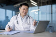 © Tetiana - Asian young male doctor consulting online, sitting in hospital office at laptop, writing in notebook and talking remotely