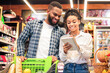 © Prostock-studio - Grocery Shopping. Black Family Couple Buying Food And Fruits Taking Notes In Checklist Choosing Groceries Together In Modern Supermarket. Spouses Buy Fresh Organic Vegetables In Store