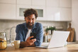 © Stockphotodirectors - A man with curly hair and a beard enjoys his coffee while looking at his smartphone with a smile in a stylish kitchen during the morning hours.