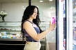 ©  Yistocking - A young Latina girl between 20 and 30 years old is shopping in a small supermarket.The woman is taking a bottle of water out of a refrigerator.Concept of small supermarkets.
