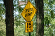 © DenisProduction.com - A bright yellow speed bump sign with a 15 MPH limit stands among green trees. The sign is clearly visible, warning drivers in a shaded, wooded neighborhood.