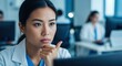 © Centric  - Thoughtful asian female doctor looking concerned while using computer in modern hospital office with colleagues working in background