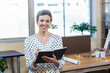 © WavebreakMediaMicro - Senior female professional standing in workspace holding black notebook and pen with sticky notes