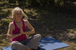 © WavebreakMediaMicro - Senior woman wearing fitness bracelet sitting on blue yoga mat in park in dappled shade meditating