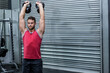 © wavebreak3 - Male athlete in red shirt lifting kettlebells at gym with weight rack and metal shutter door