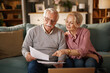© Stockphotodirectors - An elderly couple, sitting on a sofa, reviews paperwork and a laptop. The man holds the documents while the woman points at one. They appear to be planning their financial future together at home.