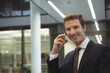 © wavebreak3 - Male business professional standing in office lobby holding smartphone and smiling, copy space